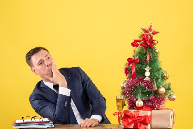 Vista frontale dell'uomo depresed che mette la mano sulla sua guancia che si siede al tavolo vicino all'albero di Natale e presenta sul giallo