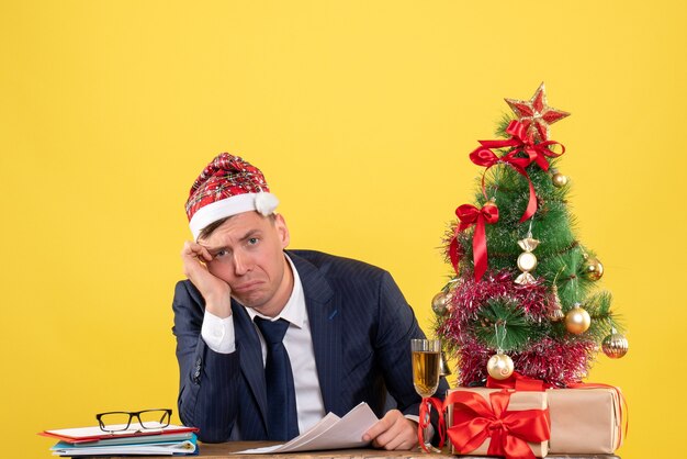 Vista frontale dell'uomo depresso con il cappello della santa che si siede al tavolo vicino all'albero di Natale e presenta su giallo.