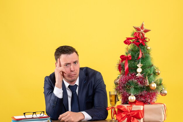 Vista frontale dell'uomo depresso seduto al tavolo vicino all'albero di Natale e regali su giallo.