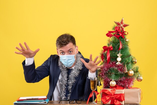 Vista frontale dell'uomo di affari che apre le sue mani che si siede al tavolo vicino all'albero di Natale e presenta su giallo