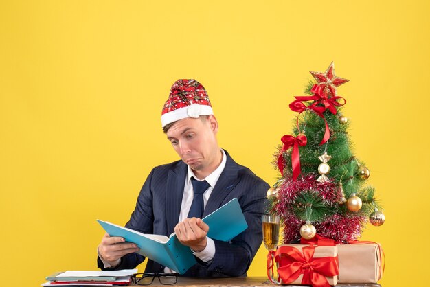 Vista frontale dell'uomo di affari che controlla i documenti che si siedono al tavolo vicino all'albero di Natale e presenta sulla parete gialla