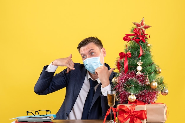 Vista frontale dell'uomo di affari che fa mi chiama il segno del telefono che si siede al tavolo vicino all'albero di Natale e presenta su giallo