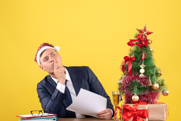 Vista frontale dell'uomo di affari che fa segno di shh che si siede al tavolo vicino all'albero di Natale e regali su giallo