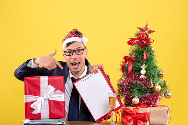 Vista frontale dell'uomo di affari che indica alla lavagna per appunti che si siede al tavolo vicino all'albero di Natale e presenta su colore giallo