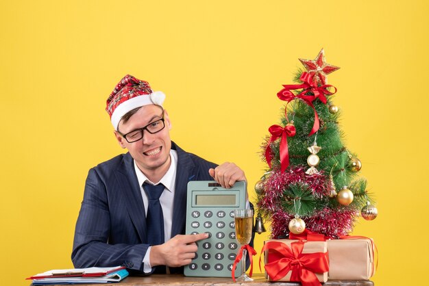 Vista frontale dell'uomo di affari che mostra la calcolatrice che si siede al tavolo vicino all'albero di Natale e presenta su giallo