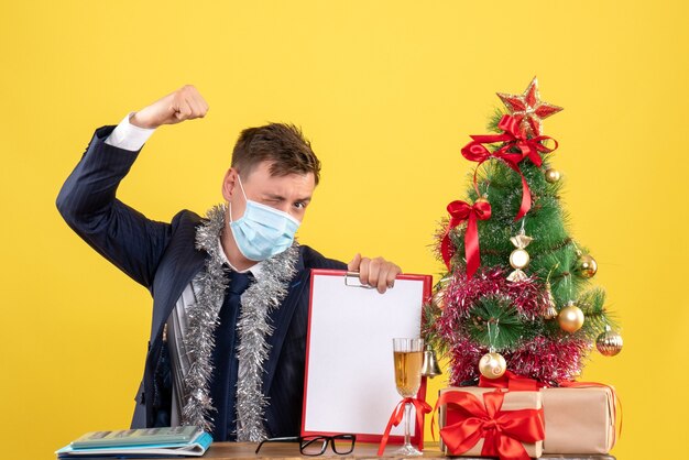 Vista frontale dell'uomo di affari che mostra la forza che si siede al tavolo vicino all'albero di Natale e presenta su giallo.