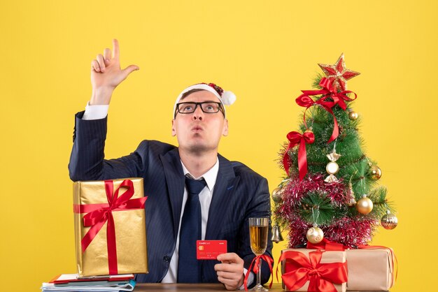 Vista frontale dell'uomo di affari che osserva in su che si siede al tavolo vicino all'albero di Natale e regali su giallo