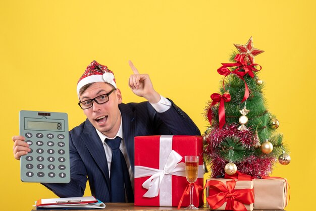 Vista frontale dell'uomo di affari che tiene l'albero di Natale del calcolatore e regali su colore giallo.