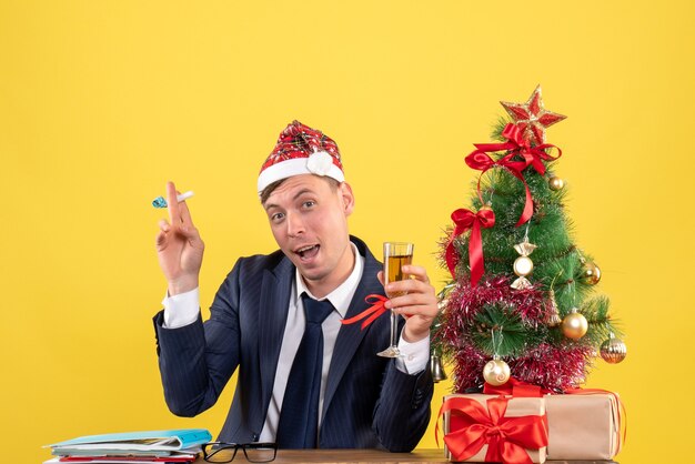 Vista frontale dell'uomo di affari che tostano il noisemaker della tenuta che si siede al tavolo vicino all'albero di Natale e presenta su colore giallo
