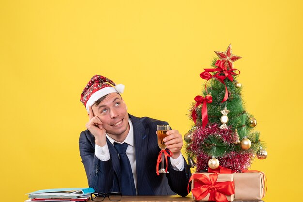 Vista frontale dell'uomo di affari che tostatura seduto al tavolo vicino all'albero di Natale e regali su giallo