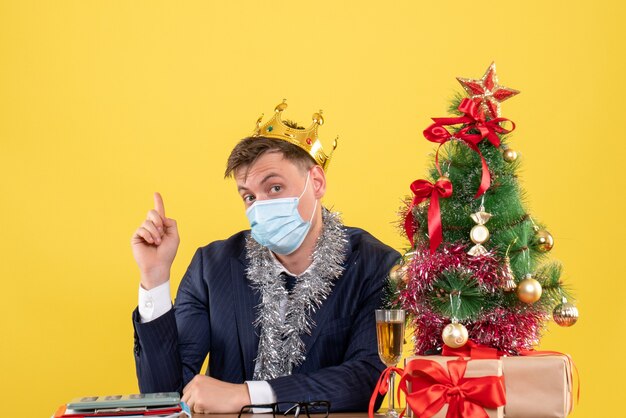 Vista frontale dell'uomo di affari con la corona che si siede al tavolo vicino all'albero di Natale e regali su giallo