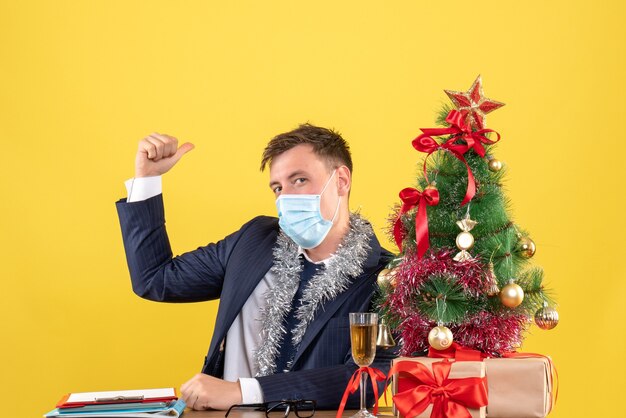 Vista frontale dell'uomo di affari con la maschera che si siede al tavolo vicino all'albero di Natale e regali su giallo