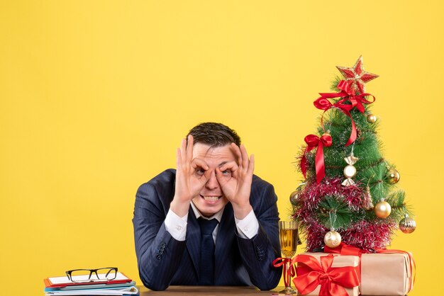 Vista frontale dell'uomo di bell'aspetto che mostra l'occhio del segno giusto che si siede al tavolo vicino all'albero di Natale e presenta su giallo