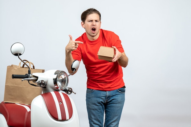 Vista frontale dell'uomo di consegna confuso in uniforme rossa che sta vicino al motorino che indica la piccola scatola su fondo bianco