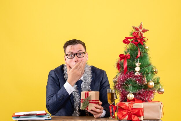 Vista frontale dell'uomo domandato che mette la mano sulla sua bocca che si siede al tavolo vicino all'albero di Natale e presenta su colore giallo