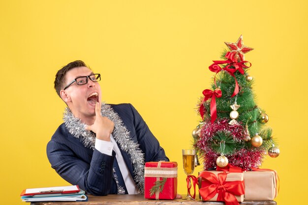 Vista frontale dell'uomo felice che apre la sua bocca che si siede al tavolo vicino all'albero di Natale e presenta su giallo