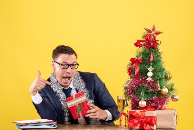 Vista frontale dell'uomo felice che indica con il dito il suo regalo seduto al tavolo vicino all'albero di Natale e regali su giallo