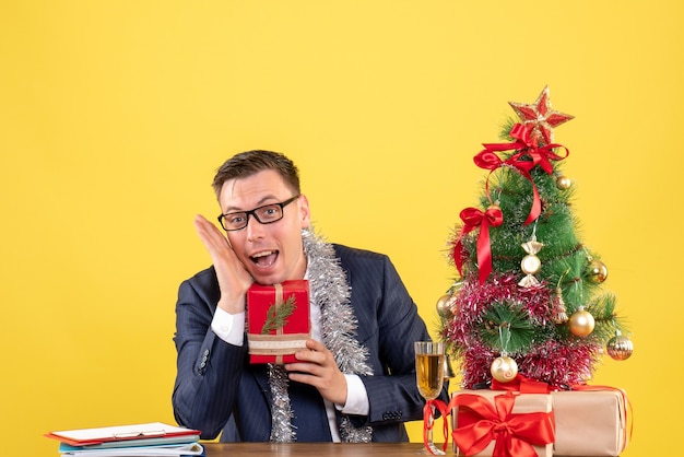 Vista frontale dell'uomo felice che tiene il regalo seduto al tavolo vicino all'albero di Natale e regali su giallo