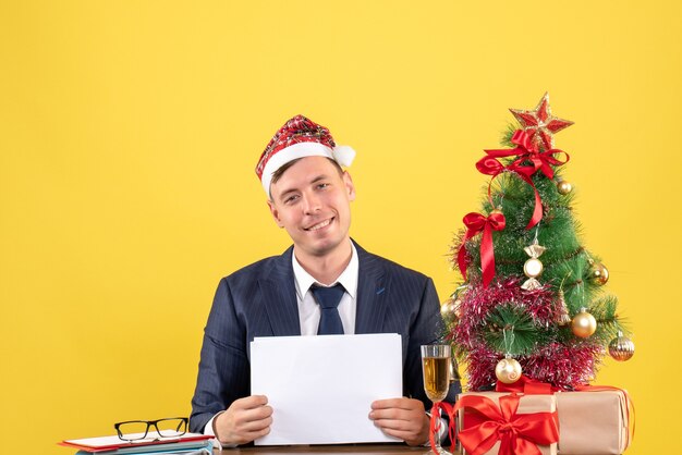 Vista frontale dell'uomo felice con il cappello della santa che si siede al tavolo vicino all'albero di Natale e presenta su giallo