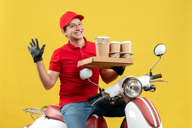 Vista frontale dell'uomo felice del corriere che indossa guanti rossi della camicetta e del cappello nella mascherina medica che trasporta l'ordine che si siede sugli ordini della tenuta dello scooter