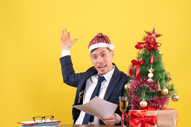 Vista frontale dell'uomo felice di affari con il cappello della Santa che si siede al tavolo vicino all'albero di Natale e regali sul giallo