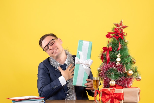 Vista frontale dell'uomo gratificato chiudendo gli occhi seduto al tavolo vicino all'albero di Natale e presenta su giallo