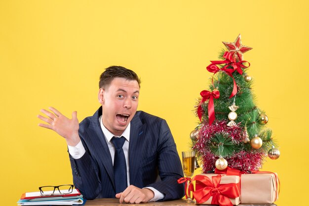 Vista frontale dell'uomo in vestito che apre la sua bocca che si siede al tavolo vicino all'albero di Natale e regali su giallo