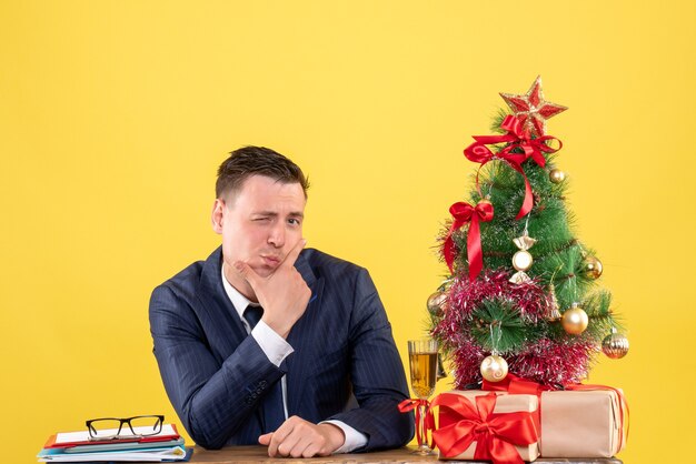 Vista frontale dell'uomo in vestito con l'occhio ammiccato che si siede al tavolo vicino all'albero di Natale e regali su giallo
