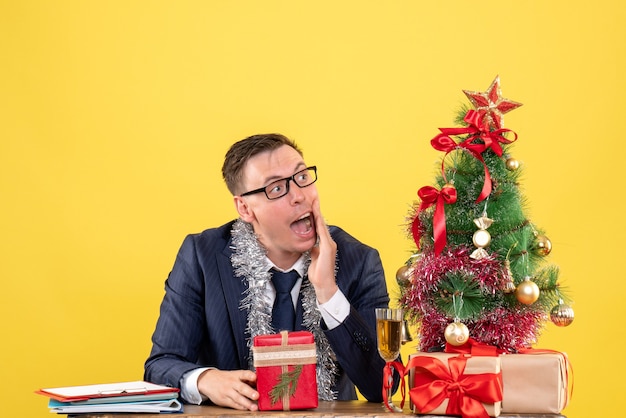 Vista frontale dell'uomo interessato guardando a destra seduto al tavolo vicino all'albero di Natale e regali su giallo