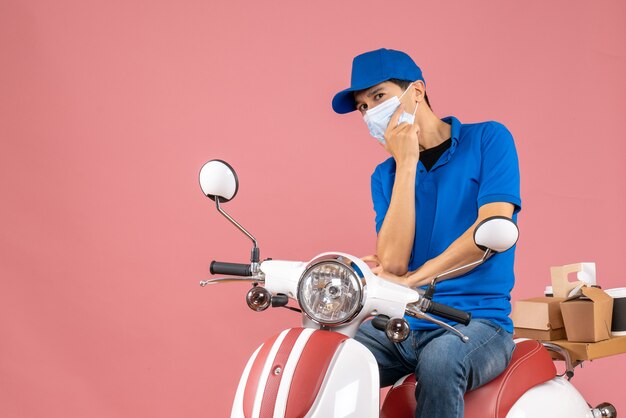 Vista frontale dell'uomo pensante corriere in maschera medica che indossa cappello seduto su scooter su sfondo pesca pastello