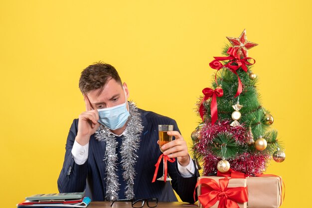 Vista frontale dell'uomo premuroso di affari che tiene champagne che si siede al tavolo vicino all'albero di Natale e regali su giallo