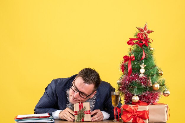 Vista frontale dell'uomo riposante che mette la testa sul giftbox seduto al tavolo vicino all'albero di Natale e regali su giallo