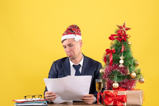 Vista frontale dell'uomo senza gioia guardando i documenti seduti al tavolo vicino all'albero di Natale e regali su giallo