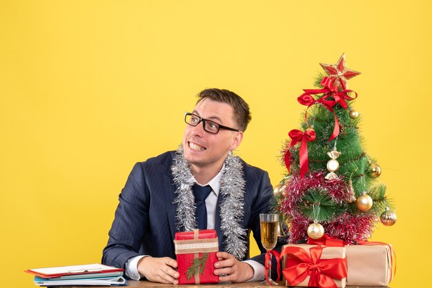 Vista frontale dell'uomo sorridente con gli occhiali seduto al tavolo vicino all'albero di Natale e regali su giallo