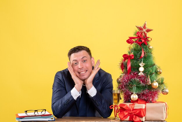 Vista frontale dell'uomo sorrise che mette le mani al suo mento che si siede al tavolo vicino all'albero di Natale e regali sul giallo