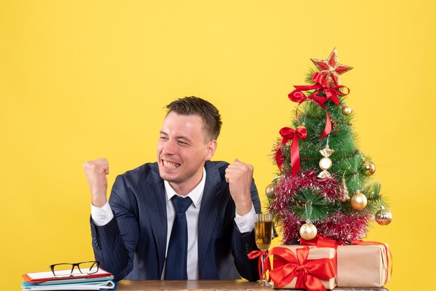 Vista frontale dell'uomo sorrise che mostra il gesto vincente seduto al tavolo vicino all'albero di Natale e regali su giallo