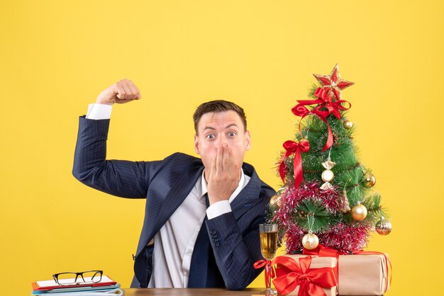 Vista frontale dell'uomo stupito che mostra il muscolo che si siede al tavolo vicino all'albero di Natale e presenta sulla parete gialla