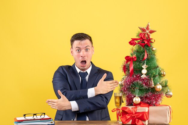 Vista frontale dell'uomo stupito che mostra le direzioni seduto al tavolo vicino all'albero di Natale e presenta sulla parete gialla