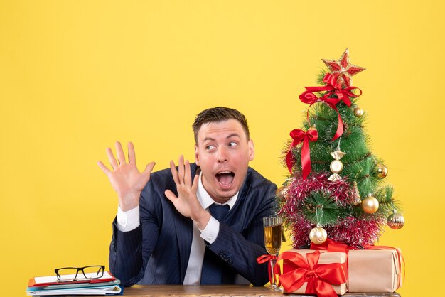 Vista frontale dell'uomo stupito in vestito che apre le sue mani seduto al tavolo vicino all'albero di Natale e regali sulla parete gialla