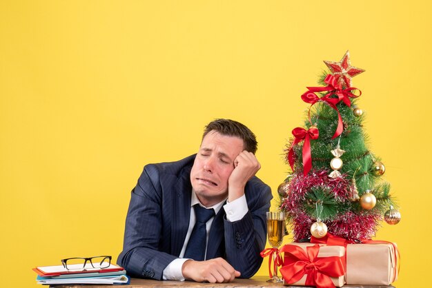 Vista frontale dell'uomo triste che si siede al tavolo vicino all'albero di Natale e regali su giallo