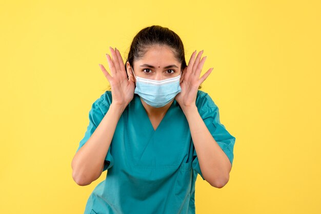 Vista frontale della bella donna medico con mascherina medica in piedi sulla parete gialla