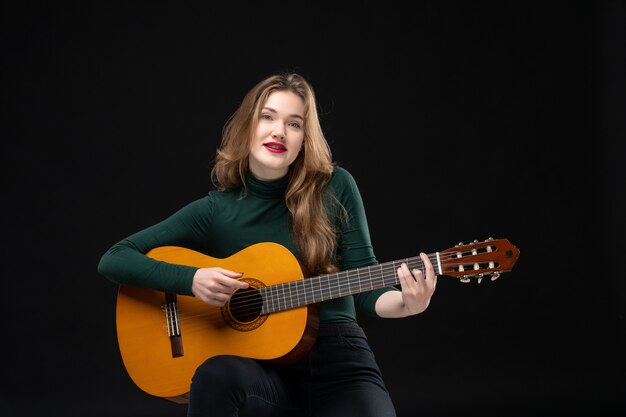 Vista frontale della bella ragazza bionda che suona la chitarra e posa per la macchina fotografica sul nero