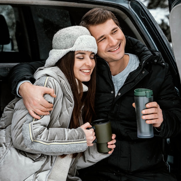Vista frontale della coppia felice con una bevanda calda nel bagagliaio dell'auto durante un viaggio su strada