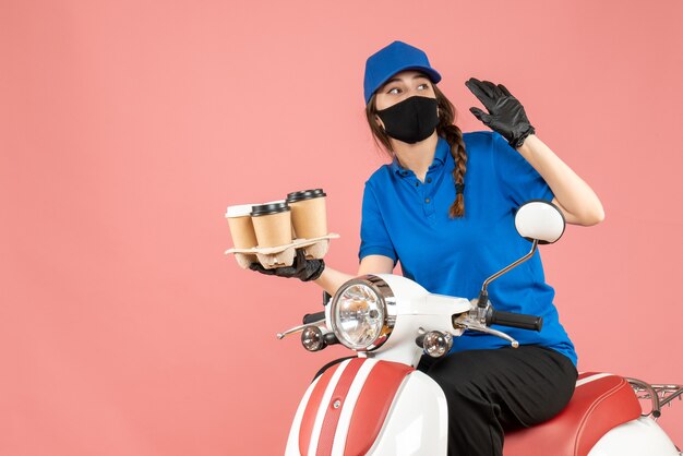 Vista frontale della curiosa donna delle consegne che indossa maschera medica e guanti seduti su uno scooter che tiene ordini su sfondo color pesca pastello pastel