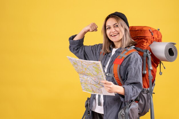 Vista frontale della donna allegra del viaggiatore con la mappa della tenuta dello zaino che mostra il muscolo del braccio