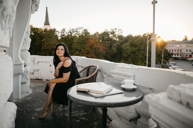 Vista frontale della donna castana attraente in vestito nero che si siede vicino al tavolino da caffè con una vista della città la sera soleggiata.