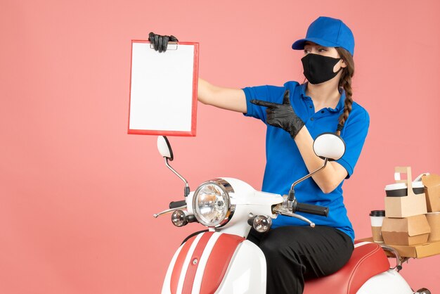 Vista frontale della donna del corriere impegnata che indossa maschera medica e guanti seduta su uno scooter con in mano fogli di carta vuoti che consegnano ordini su sfondo color pesca pastello