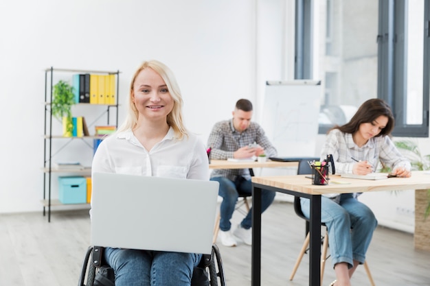Vista frontale della donna di smiley in sedia a rotelle che posa con il computer portatile sul lavoro