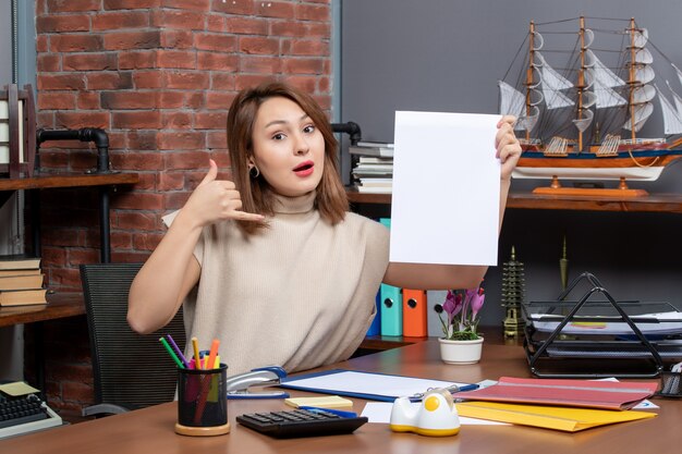 Vista frontale della donna meravigliata che mi fa chiamare il segno del telefono seduto in ufficio