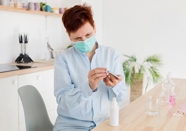 Vista frontale della donna più anziana che disinfetta il suo smartphone mentre indossa maschera medica